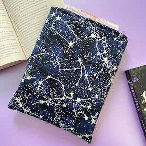 May include: A navy blue book sleeve with a white constellation design. The text "BEFORE SHE WAS DEADLY. NOW SHE IS UNSTOPPABLE" is printed on the sleeve. The sleeve is protecting a book.
