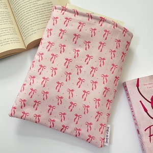 May include: A pink book sleeve featuring a pattern of red bows. The sleeve is open, revealing a book's pages. A book with a pink cover is visible on the right. The text "Snug as a Book" is on the bottom right.