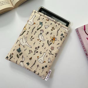 May include: Beige fabric tablet sleeve featuring a pattern of geese, flowers, and foliage. The sleeve, designed for a tablet, has a tag reading "Snug as a Book". A book and open book are visible in the background.