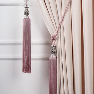 Może przedstawiać: Frędzle do zasłon w kolorze brudnego różu ze srebrnymi elementami dekoracyjnymi. Frędzle wiszą na różowym sznurku i znajdują się obok jasnoróżowej zasłony. Ściana jest biała z ozdobnymi listwami.