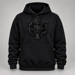 May include: A black hooded sweatshirt featuring a large, dark, textured bear head graphic on the chest. The hoodie includes a front kangaroo pocket, long sleeves with ribbed cuffs, a matching ribbed hem, and black drawstring cords at the neckline.