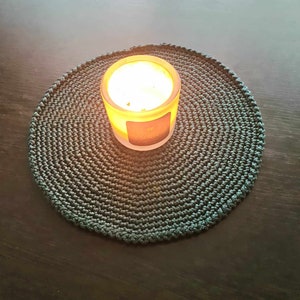 May include: A teal crocheted coaster with a round shape and a lit candle in the center.