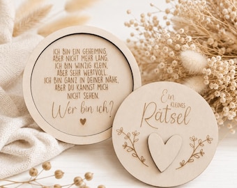 May include: Two circular light-colored wooden plaques. The larger plaque features engraved German text: "Ich bin ein Geheimnis, aber nicht mehr lang. Ich bin winzig klein, aber sehr wertvoll. Ich bin ganz in deiner Nähe, aber du kannst mich nicht sehen. Wer bin ich?" The smaller plaque displays the text "Ein kleines Rätsel" with a wooden heart cutout and floral engravings.