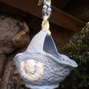 May include: A light blue ceramic hanging basket with a white cameo design. The basket is decorated with floral details and has a rope handle with blue and white beads.