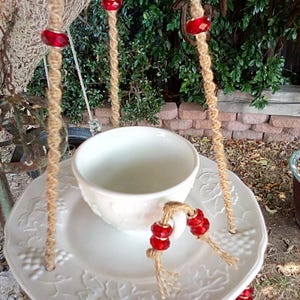 May include: A white porcelain teacup and saucer hanging from a rope with red beads. The saucer has a grapevine design.