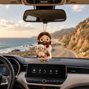 May include: A crocheted hanging ornament featuring a figure with brown hair and a beard, wearing a cream-colored robe with a red sash, holding a small white lamb. The ornament is suspended by a tan string from a vehicle rearview mirror.