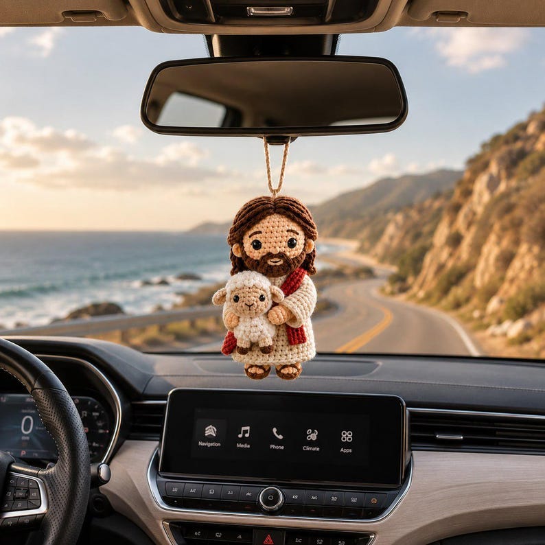 May include: A crocheted hanging ornament featuring a figure with brown hair and a beard, wearing a cream-colored robe with a red sash, holding a small white lamb. The ornament is suspended by a tan string from a vehicle rearview mirror.