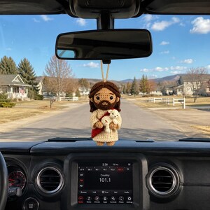 May include: A crocheted hanging ornament featuring a figure with brown hair and a beard, wearing a cream-colored robe with a red sash, holding a small white lamb. The figure is suspended by a tan string from a vehicle rearview mirror.