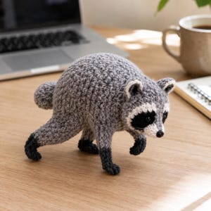 May include: A crocheted raccoon figurine crafted from grey yarn with a white face patch, black eye markings, and dark grey paws. The small stuffed animal features a rounded body, a short tail, and a black nose, standing in a forward-leaning pose.