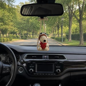 May include: A crocheted Snoopy figure wearing a brown aviator cap and goggles, holding a red heart, suspended from a beige yarn swing. The ornament hangs from the rearview mirror of a vehicle, featuring a white body and black ears.