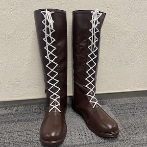 Puede incluir: Un par de botas de cuero marrón de altura por encima de la rodilla con cordones blancos. Los cordones están atados en forma de cruz en la parte delantera de las botas.