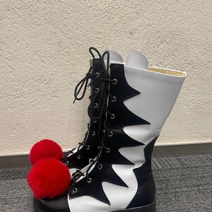 Puede incluir: Botas altas blancas y negras con pompones rojos en los dedos. Las botas tienen un patr&oacute;n blanco y negro irregular y cordones negros.