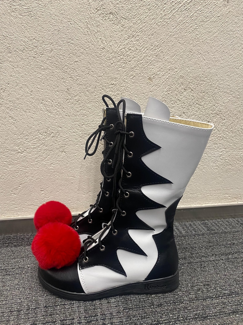 Puede incluir: Botas altas blancas y negras con pompones rojos en los dedos. Las botas tienen un patr&oacute;n blanco y negro irregular y cordones negros.
