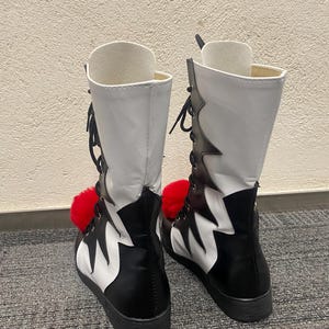 Puede incluir: Un par de botas blancas y negras con pompones rojos. Las botas tienen un dise&ntilde;o dentado y est&aacute;n atadas en la parte delantera.