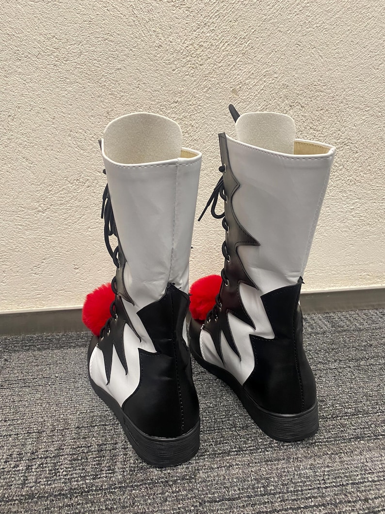 Puede incluir: Un par de botas blancas y negras con pompones rojos. Las botas tienen un dise&ntilde;o dentado y est&aacute;n atadas en la parte delantera.