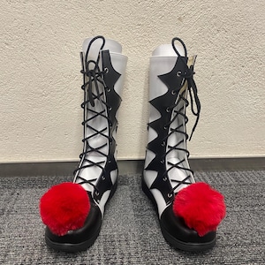 Puede incluir: Un par de botas blancas y negras con pompones rojos en los dedos. Las botas tienen un cierre de cordones en la parte delantera y una punta puntiaguda.