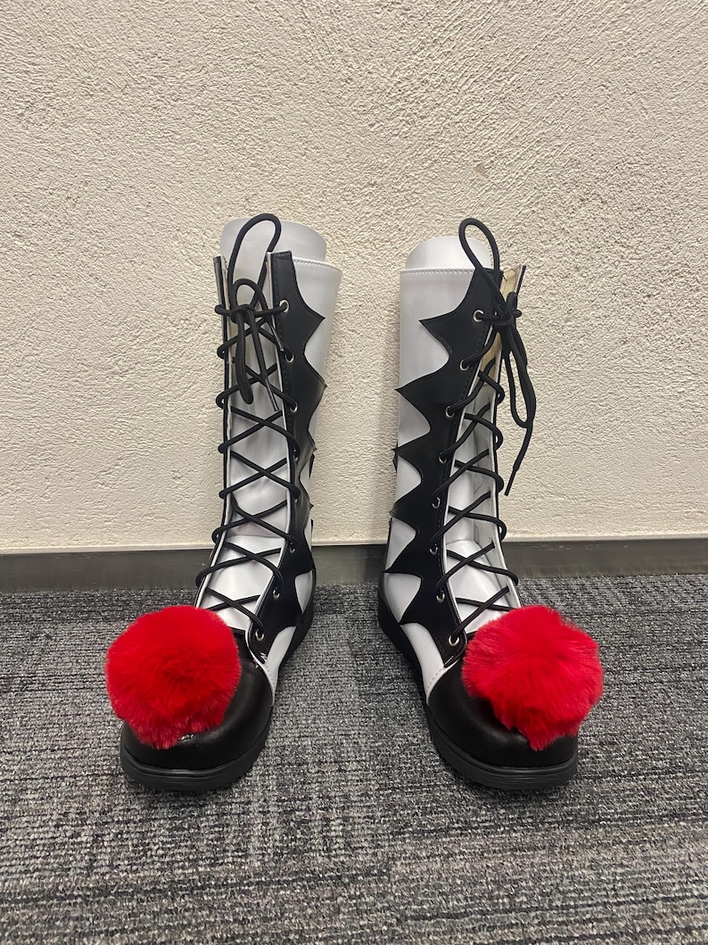 Puede incluir: Un par de botas blancas y negras con pompones rojos en los dedos. Las botas tienen un cierre de cordones en la parte delantera y una punta puntiaguda.
