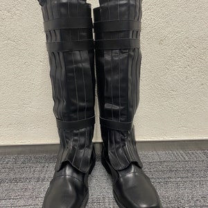 May include: A pair of black knee-high boots. The boots have a smooth, matte finish with vertical pleats and two straps. The toe is rounded and the sole is flat.