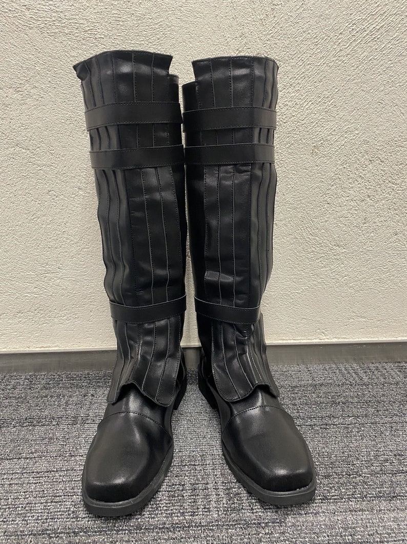 May include: A pair of black knee-high boots. The boots have a smooth, matte finish with vertical pleats and two straps. The toe is rounded and the sole is flat.