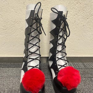 Puede incluir: Botas negras y blancas de ca&ntilde;a alta con pompones rojos en los dedos. Las botas tienen un cierre de cordones en la parte delantera y una punta puntiaguda.