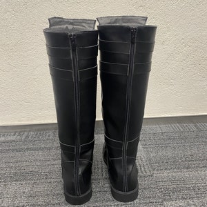 May include: A pair of black knee-high boots with zippers running down the back. The boots have multiple straps and buckles for a decorative touch. The boots are made of a smooth, leather-like material.