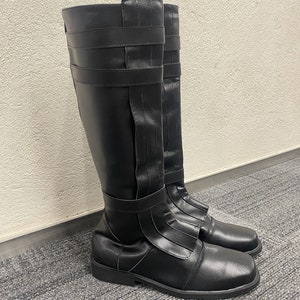 May include: A pair of black knee-high boots. The boots have a smooth, matte finish and feature multiple straps and buckles. The design includes a square toe and a low, flat heel. The boots are made of a synthetic material.