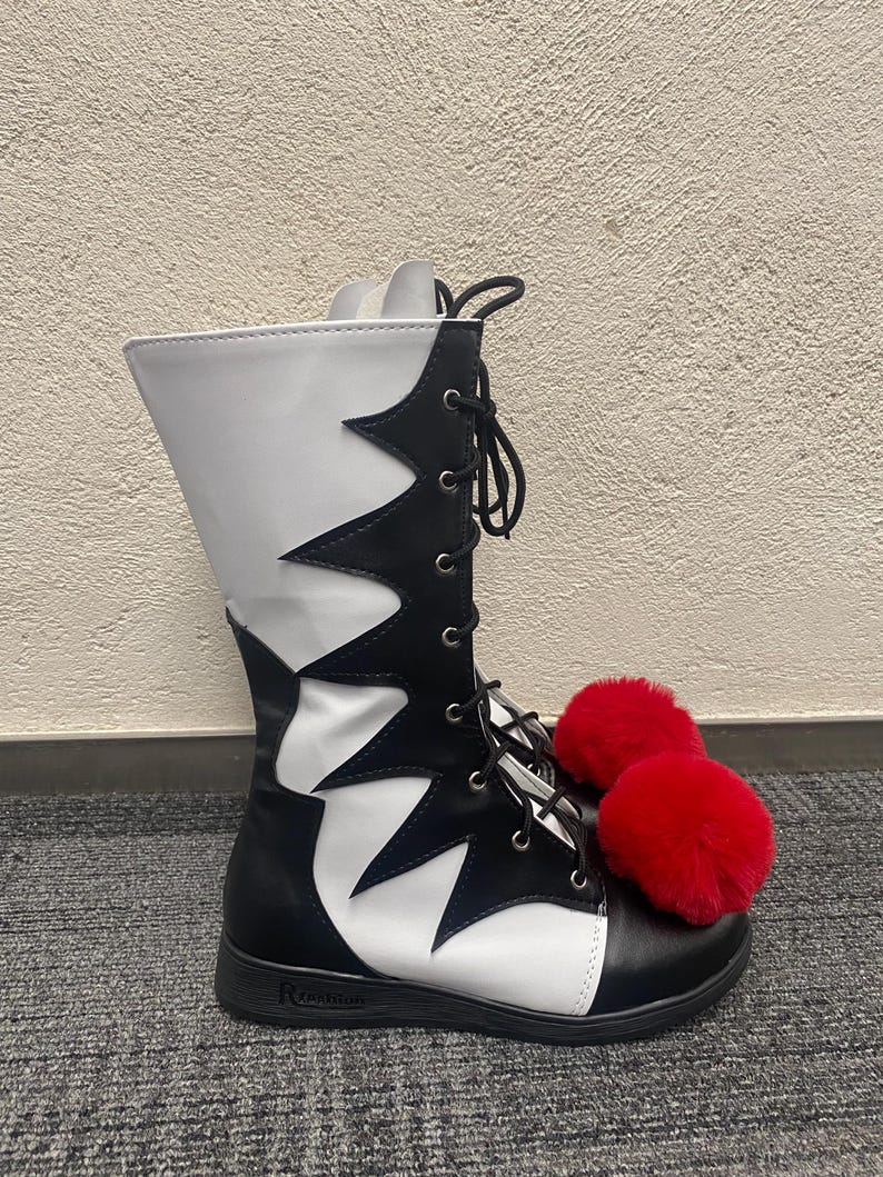 Puede incluir: Botas negras y blancas de ca&ntilde;a alta con pompones rojos peludos. Las botas tienen un cierre de cordones en la parte delantera y una suela blanca.