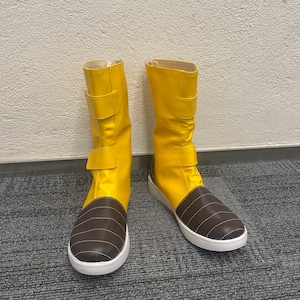 Puede incluir: Un par de botas amarillas con detalles de cuero marrón y suelas blancas. Las botas tienen una cremallera en el lateral y una correa con hebilla en la parte superior.