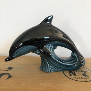 Könnte beinhalten: Eine schwarze und blaue Keramik-Delfinfigur, die aus einer Welle springt. Der Delfin hat eine glänzende Oberfläche und befindet sich in einer Sprungpose. Die Basis der Figur ist eine Welle mit einer blauen und grünen Glasur.