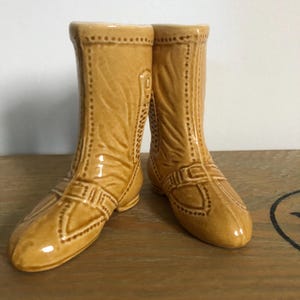 May include: A pair of ceramic boot salt and pepper shakers. The shakers are a light brown color and have a detailed design.
