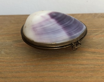 May include: A decorative clam shell box with a hinged lid. The shell is white and purple, set in a brass-colored metal frame. The box is resting on a wooden surface.