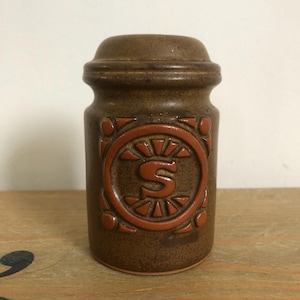 Puede incluir: Un salero de cerámica marrón con tapa marrón y un círculo rojo con la letra "S" en el centro.