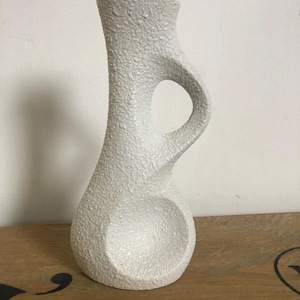 May include: White ceramic vase with a textured surface and a unique, curved design. The vase has a cutout handle and a wide opening.