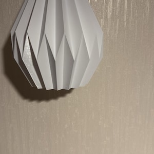 Puede incluir: Lámpara colgante geométrica blanca con cable negro y montaje en el techo. La pantalla tiene un diseño facetado, tipo origami. La luz cuelga contra una pared texturizada de tono neutro.