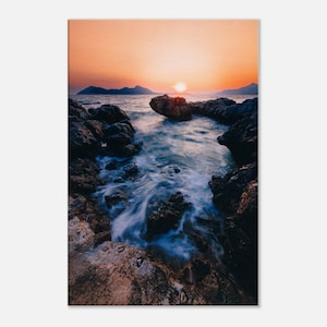 Puede incluir: Una puesta de sol pintoresca sobre una costa rocosa con el sol poniéndose detrás de las montañas en la distancia. El agua es de un azul profundo y las rocas son de un gris oscuro. El cielo es de un naranja y un rosa vibrantes.