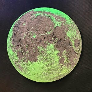 May include: A close-up of a round, grey and green moon-shaped object. The object is textured and has a glow-in-the-dark effect.