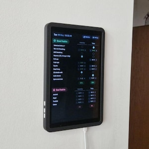 Puede incluir: Una tableta negra que muestra un rastreador de hábitos, montada en una pared blanca. La pantalla muestra una lista de buenos y malos hábitos con indicadores de progreso. La tableta está en una funda negra y está conectada a un cable de carga blanco.