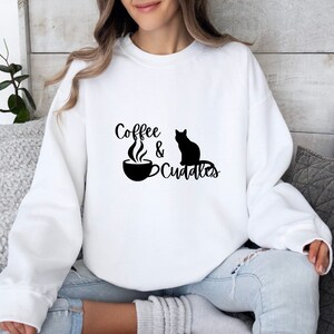 Może przedstawiać: Biała bluza z czarnym napisem "Coffee & Cuddles" i czarną sylwetką kota.