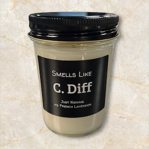 May include: A clear glass jar candle with a black lid. The candle has a black label with the text "Smells Like C. Diff" and "Just Kidding Its French Lavender". The candle is filled with a light beige wax.