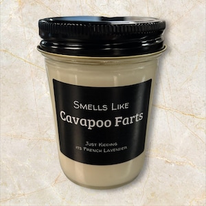 May include: A glass jar candle with a black lid and a black label. The label reads "Smells Like Cavapoo Farts" with the text "Just Kidding ITS FRENCH LAVENDER" below. The candle has a light beige color.