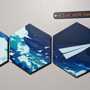 May include: Three hexagonal canvas paintings with a blue and white sky background. The left two paintings feature white paper airplanes flying through the sky. The rightmost painting features a single, larger paper airplane flying through the sky. The paintings are 8.5 inches wide and 9.8 inches tall.