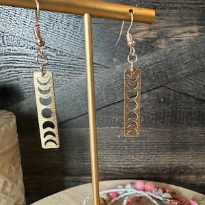 May include: Gold tone earrings with a crescent moon design. The earrings are hanging from a gold stand.