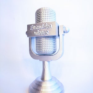 Op de afbeelding: Zilverkleurige microfoontrofee met de tekst "Eurovision Winner". De microfoon heeft een afgeronde bovenkant en een cirkelvormige basis. De trofee is gemaakt van een metaalachtig materiaal.