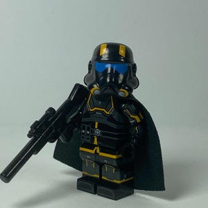 May include: A black and yellow action figure of a character in a helmet with blue eye lenses, holding a black rifle. The figure has a black cape and yellow accents on the armour. The helmet has yellow stripes.