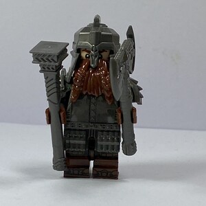 May include: A grey Lego minifigure of a dwarf warrior with a red beard, wearing a helmet with a pointed crest, holding a grey axe and a grey staff.
