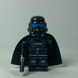 May include: A black and blue action figure of a character in a helmet and cape, holding a black rifle. The figure has blue lenses and a dark blue and grey uniform. The cape is black.