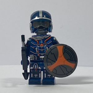 May include: A blue and grey Lego minifigure with a black gun and a grey and orange shield. The minifigure is wearing a blue helmet with a silver stripe.