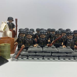 May include: A group of 18 miniature plastic toy soldiers wearing black and grey camouflage uniforms and helmets. The soldiers are holding rifles and are standing on a grey base with sandbags. There is a white horse and a soldier on horseback in the background.