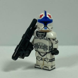 May include: A white and blue Lego minifigure of a clone trooper holding a black blaster rifle. The helmet has blue and red markings. The figure has detailed armour with grey and black accents. The background is a plain, light grey.