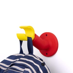 Puede incluir: Un gancho de pared rojo y amarillo con una mochila a rayas azules y blancas colgando. El gancho está diseñado con una base circular y un brazo amarillo curvado. La mochila tiene un patrón de rayas.